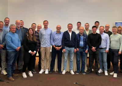 Gruppenfoto von etwa 25 Personen, die Mitglieder des Landesbeirats Holz Berlin-Brandenburg sind. Sie stehen in mehreren Reihen in einem Konferenzraum vor einer weißen Wand. Einige Personen tragen Freizeitkleidung, andere Business Casual. Es sind sowohl Männer als auch Frauen vertreten. Rechts im Bild befindet sich ein Roll-Up mit Logos von Partnerunternehmen des Holzbauclusters Berlin-Brandenburg. Die Stimmung wirkt freundlich und professionell.