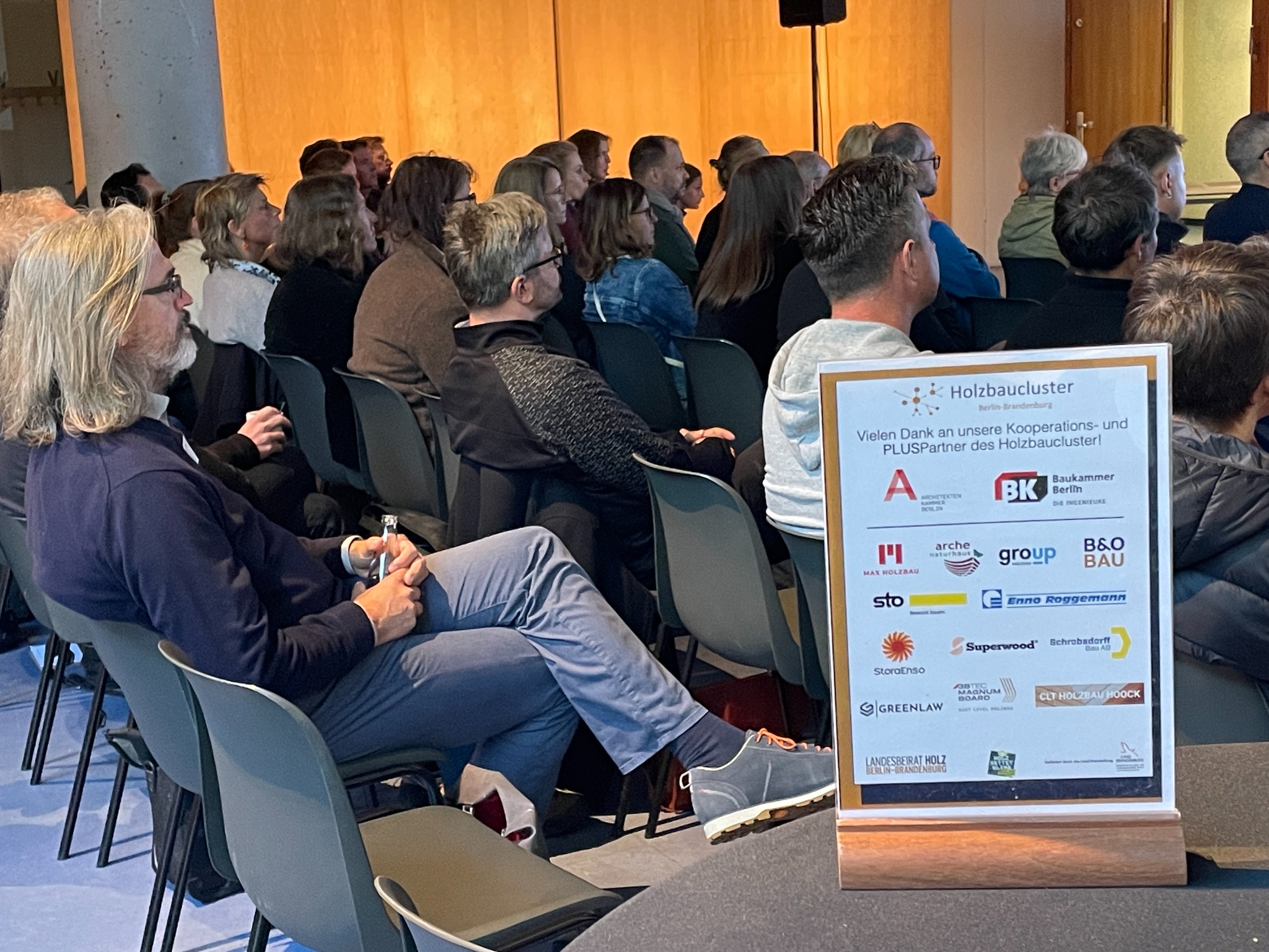 Teilnehmende eines Holzbaucluster-Seminars mit Aufsteller der PLUS-Partner im Vordergrund